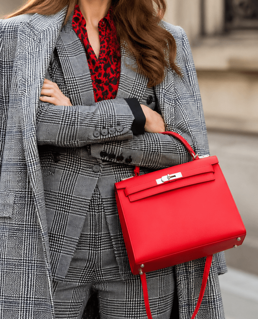 A woman in a grey check suit holding a red Hermès Kelly bag